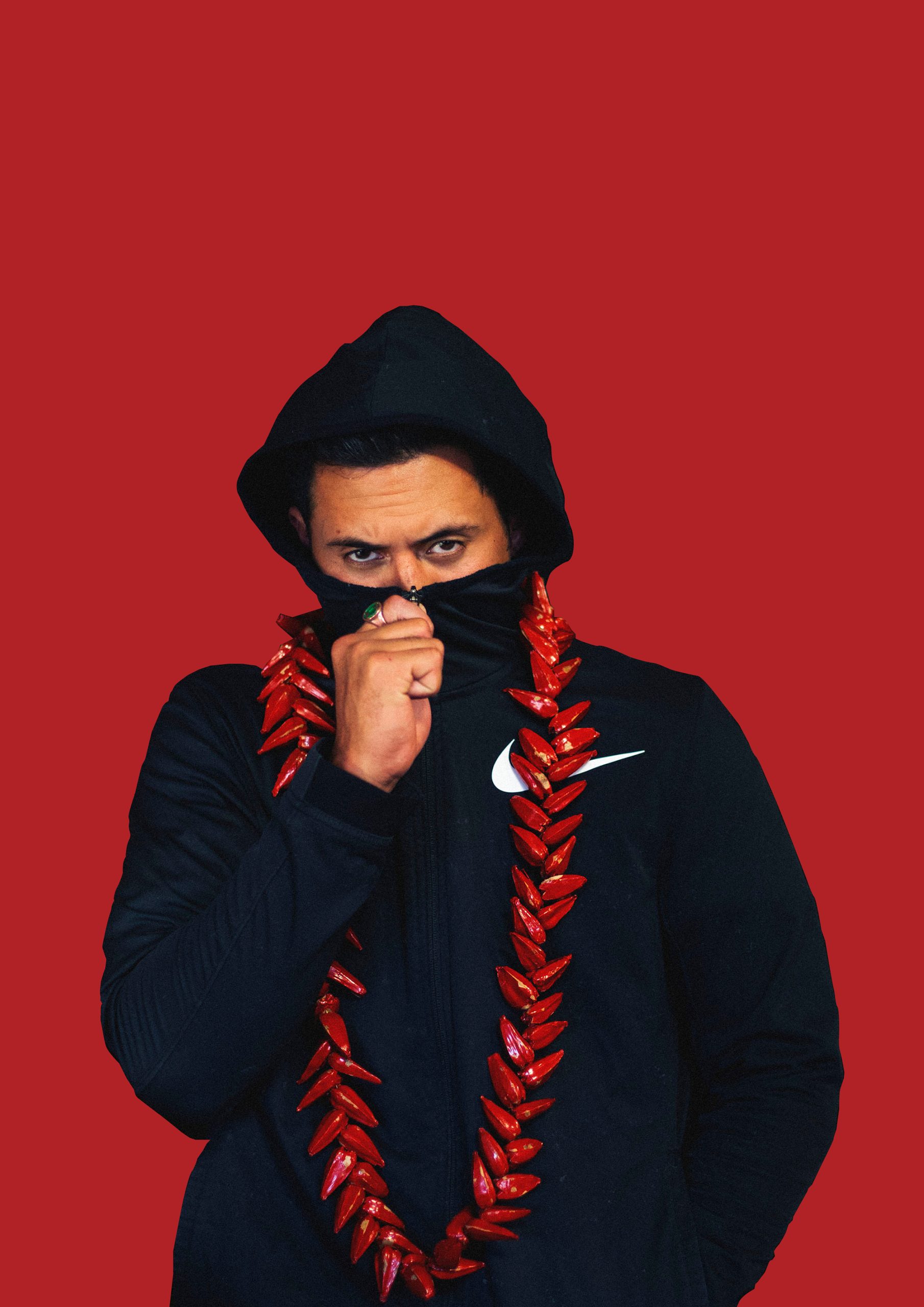 We see a man against a bright red background wearing a black Nike hoodie. He is holding the hoodie to his nose and is wearing a large necklace made out of chilles around his neck.